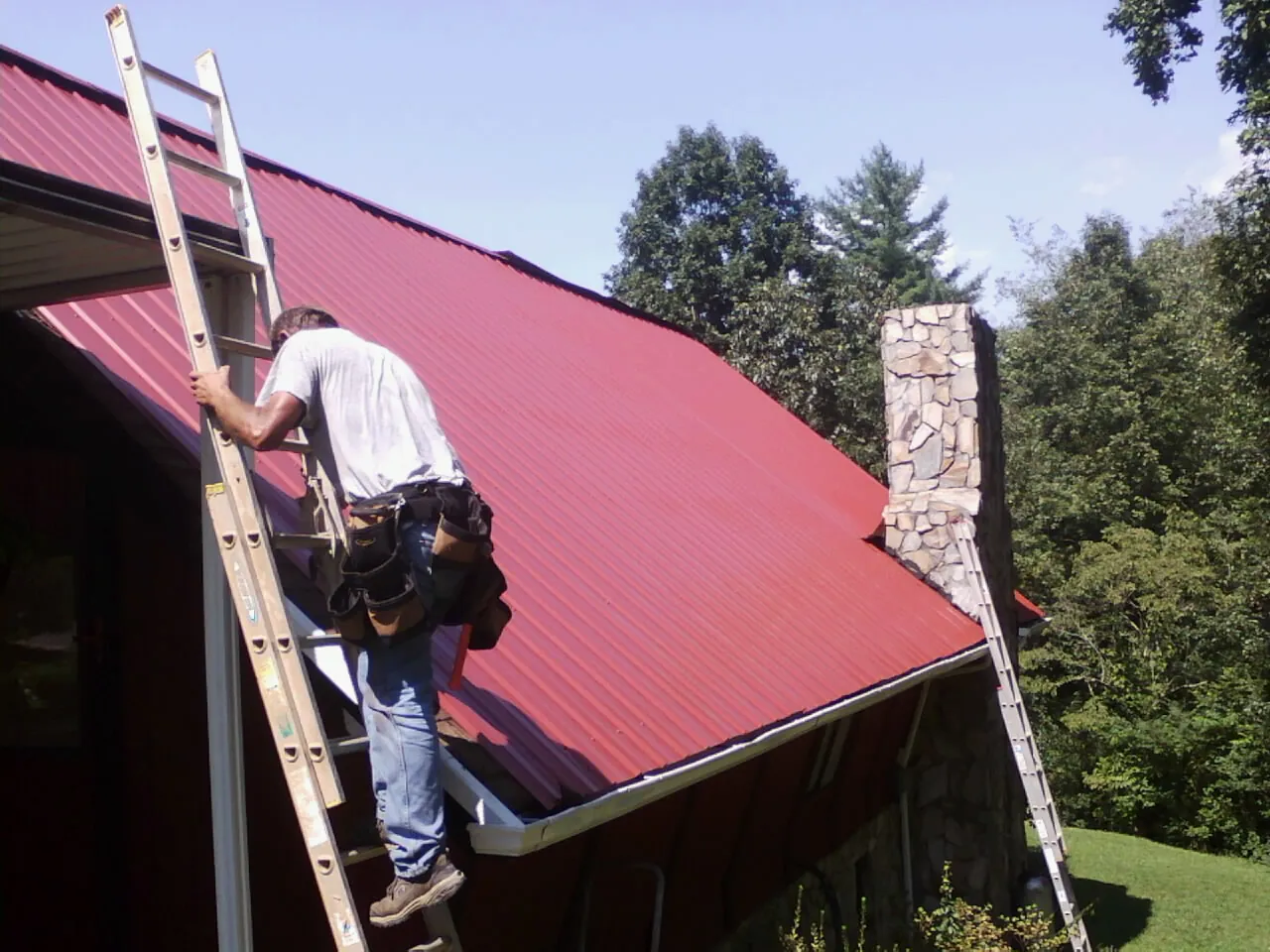 Roofer on a ladder beside a red metal roof and stone chimney — Soffit & Fascia Repair by Roofing Brothers in Rock Creek